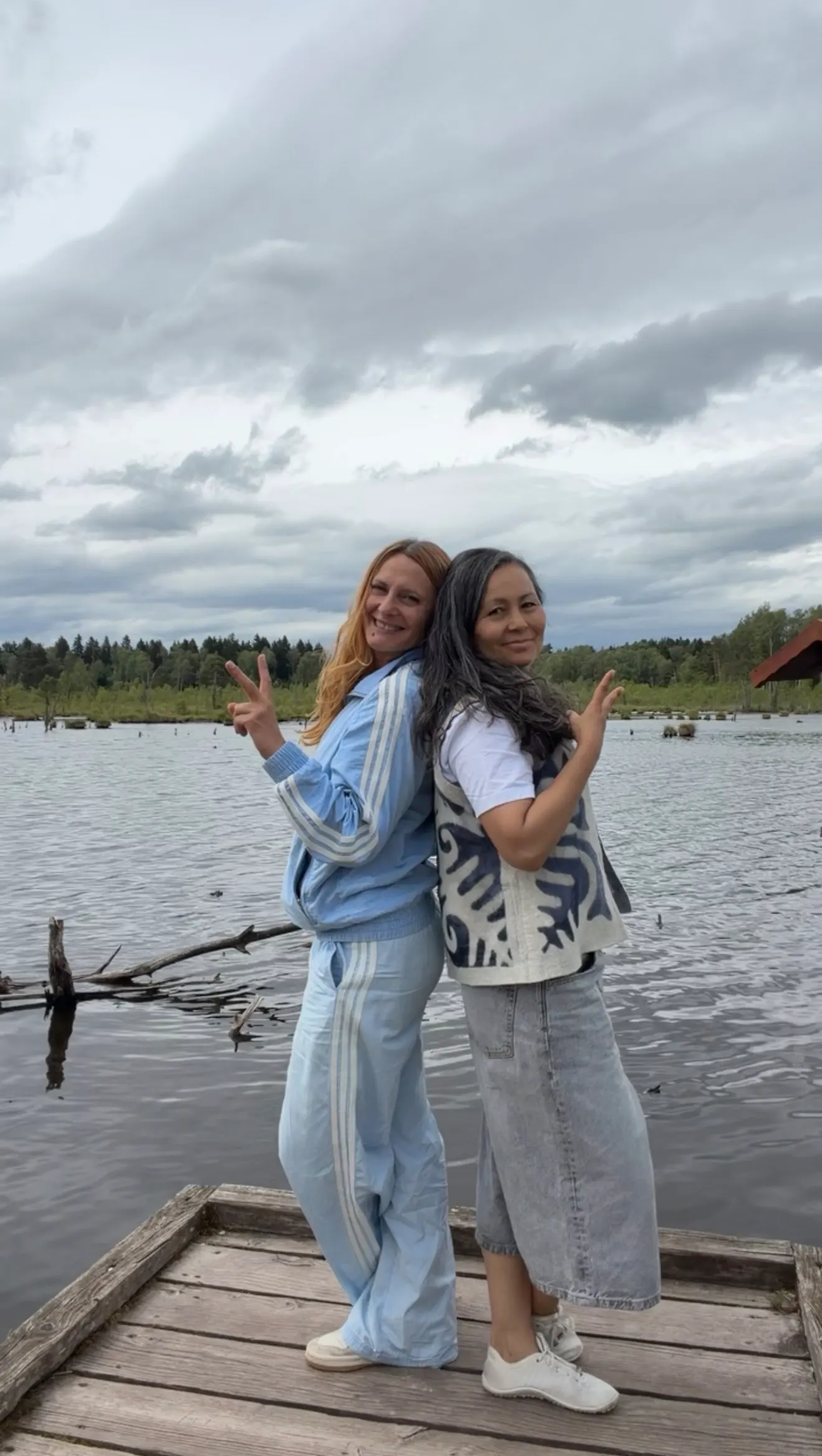 Sonia und Jeanna stehen lachend Rücken an Rücken auf einem Holzsteg am See